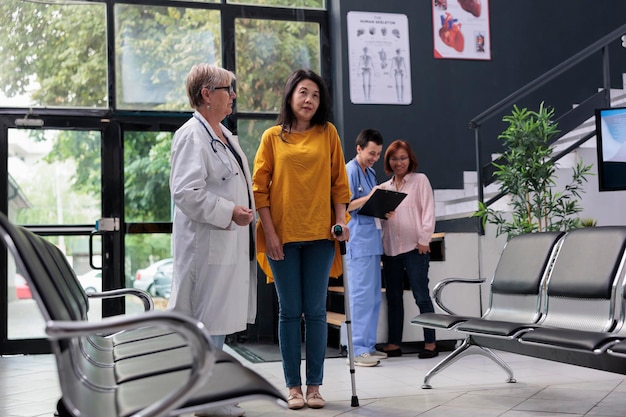 Senior physician helping injured patient to walk with stick, suffering from physical impairment and using cane. Doctor attending rehabilitation therapy to recover from leg injury at facility.