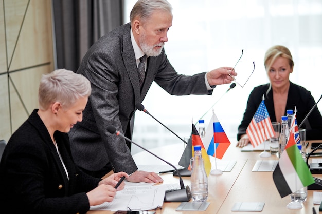 Senior politician offers his plan of action and share opinion, elderly man in suit talking to multi-ethnic group of partners sitting at desk in boardroom, discussing