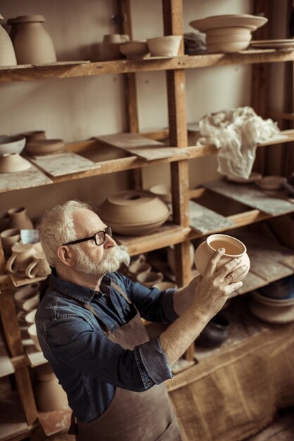 Senior potter in apron and eyeglasses examining ceramic bowl at workshop