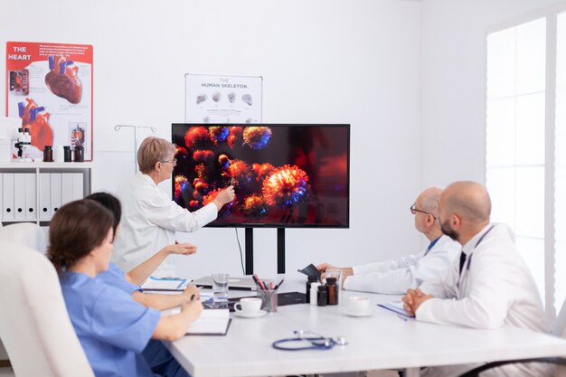 Senior practitioner doctor woman pointing virus sickness on monitor in hospital meeting room discussing with coworkers during brainstorming