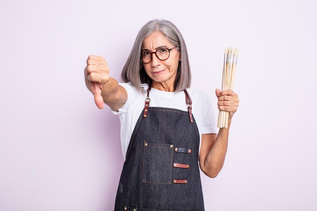 Senior pretty woman feeling cross,showing thumbs down. artist with brushes