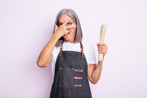 Senior pretty woman looking shocked scared or terrified covering face with hand artist with brushes