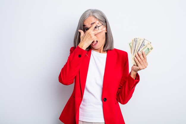 Senior pretty woman looking shocked, scared or terrified, covering face with hand. business and banknotes concept