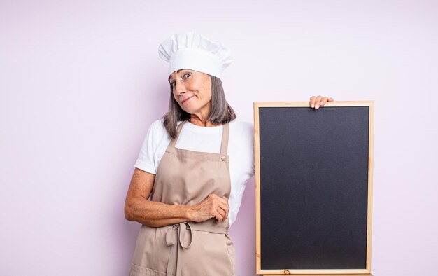 senior pretty woman shrugging, feeling confused and uncertain. chef and blackboard concept