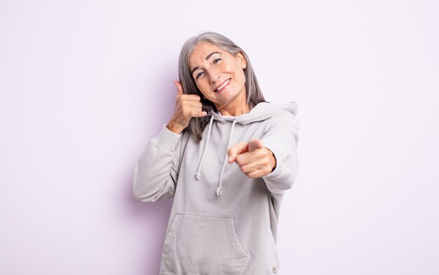 Senior pretty woman smiling cheerfully and pointing to camera while making a call you later gesture, talking on phone