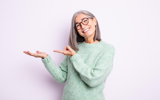 Senior pretty woman smiling cheerfully and pointing to copy space on palm on the side, showing or advertising an object