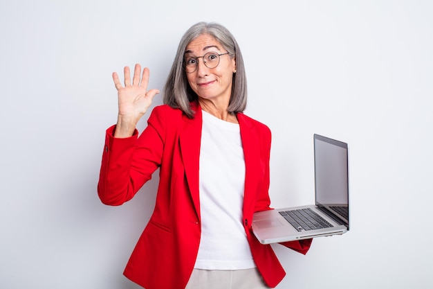 Senior pretty woman smiling happily, waving hand, welcoming and greeting you. business and laptop concept