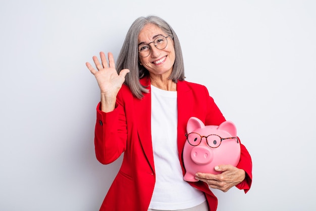 Senior pretty woman smiling happily, waving hand, welcoming and greeting you. piggy bank concept