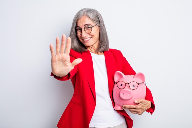 Senior pretty woman smiling and looking friendly, showing number five. piggy bank concept