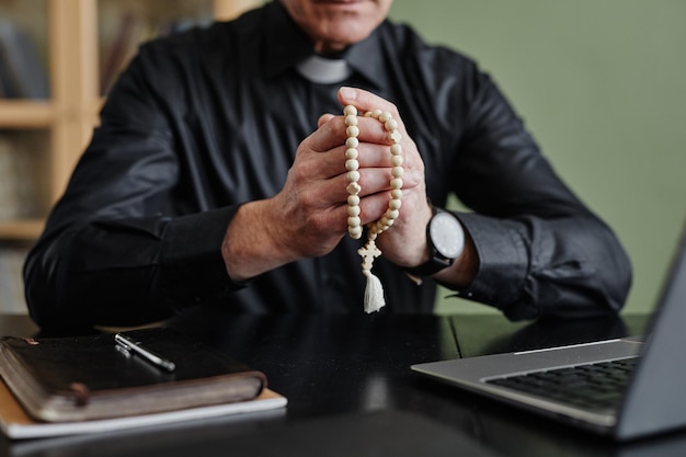 Senior priest holding rosary praying at desk in office