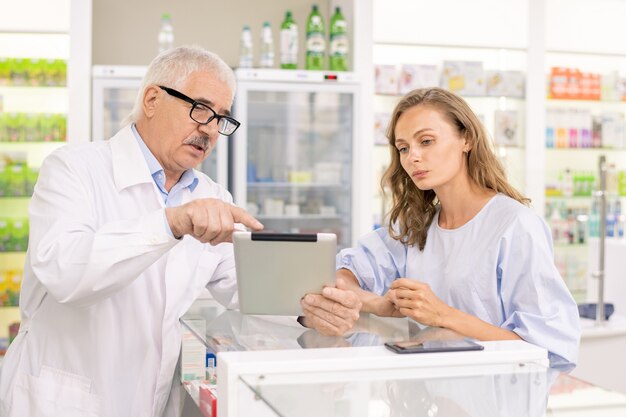 Senior professional worker of contemporary drugstore showing his young female trainee online information about new anti-covid19 medicine