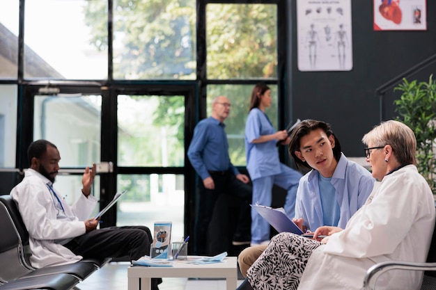 Senior pyhysician medic standing beside asian patient helping to fill insurance document before start examination during checkup visit consultation in hospital reception. Health care service and conce