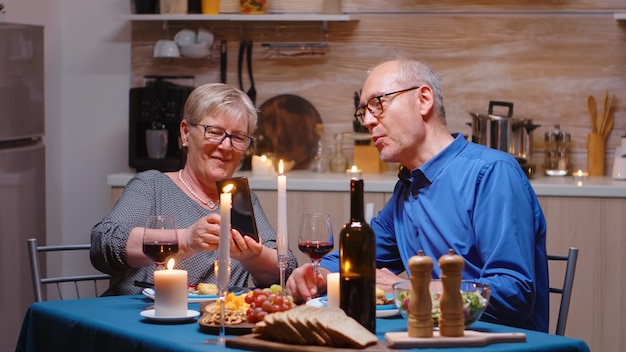 Anziana moglie in pensione che mostra le foto sullo smartphone al marito durante una cena romantica. coppia seduta al tavolo, parlando, usando il telefono, celebrando il loro anniversario nella sala da pranzo.