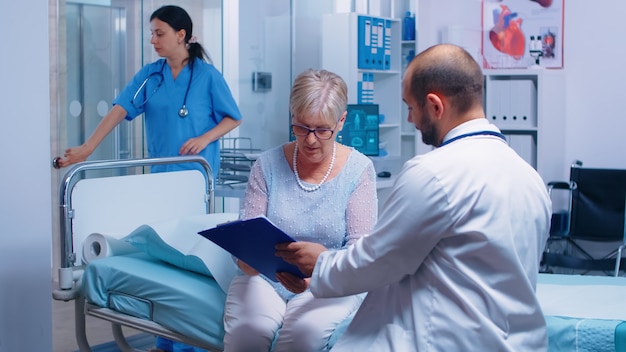 Senior donna in pensione firma per il rilascio medico nella moderna clinica privata seduta sul letto d'ospedale, infermiera che lavora in background. paziente medico di consultazione di medicina di sanità