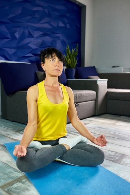 Senior rijpe vrouw, zittend in lotushouding op yoga mat, stretching oefening in de huiskamer. Gezond levensstijlconcept.