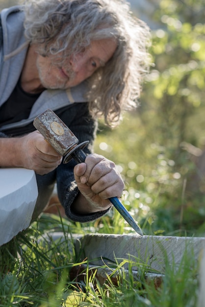 Senior sculptor sculpting with chisel and hammer in stone outdoors.