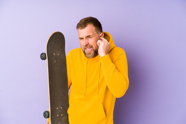 Senior skater man isolated on purple wall covering ears with hands.