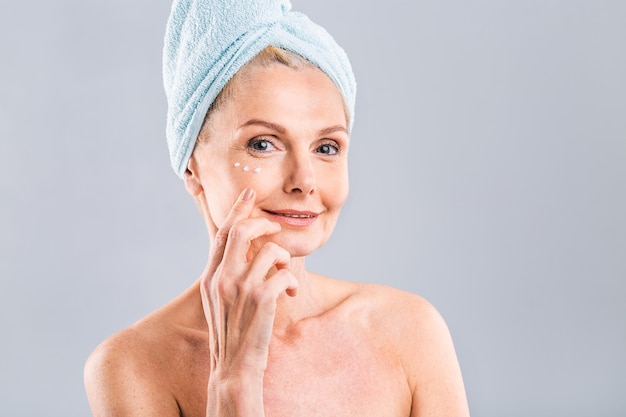 senior smiling 50s middle aged mature older woman applying facial cream on face looking at camera anti age healthy dry skin care beauty therapy concept old skincare treatment