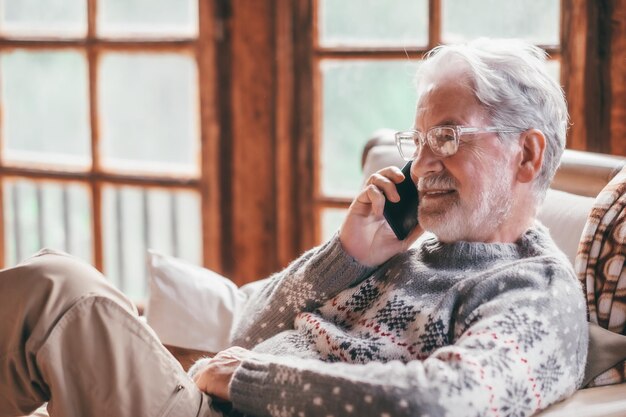 수염이 흰 머리를 가진 웃고 있는 노인이 휴대전화를 사용하여 안락의자에 편안하게 집에 앉아 있습니다.