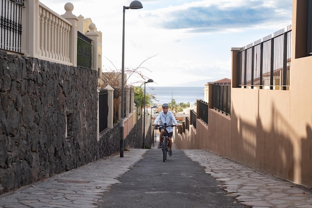 Senior smiling woman cycling with her electric bicycle in uphill alley Horizon over water in background