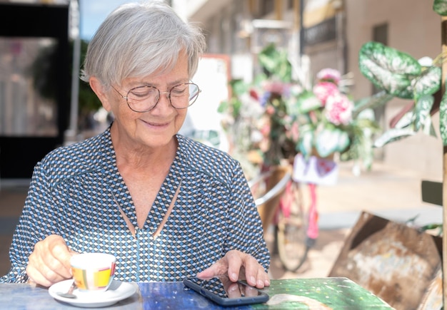 Пожилая улыбающаяся женщина сидит в уличном кафе со своим смартфоном, держа в руке чашку кофе