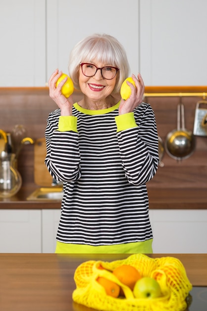 senior smiling woman in striped sweater holding lemons for lemonade while standing in the kitchen