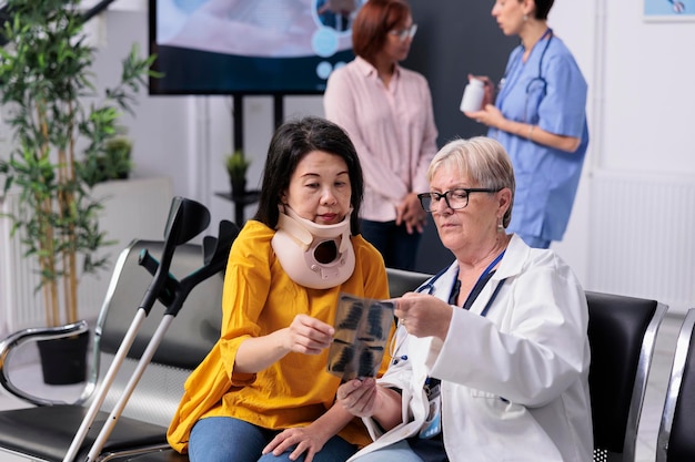 Senior specialist consulting patient with cervical neck collar looking at medical xray exam during checkup visit in hospital waiting room. Asian woman with physical pain and injury having appointment.