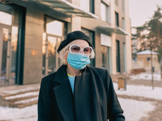 senior stijlvolle vrouw in baret en elegante zwarte jas en met medisch masker buiten wandelen