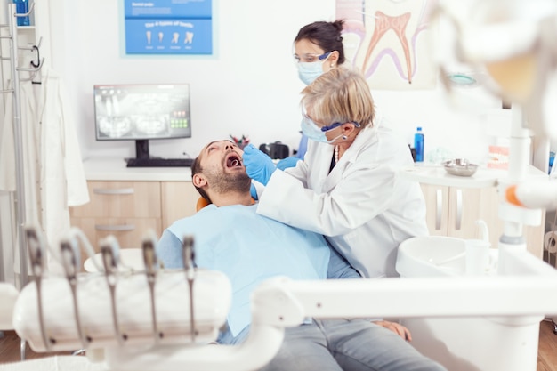 Photo senior stomatologist woman with protection face mask performing dental inspection after putting implant to sick man mouth in orthodontic clinic