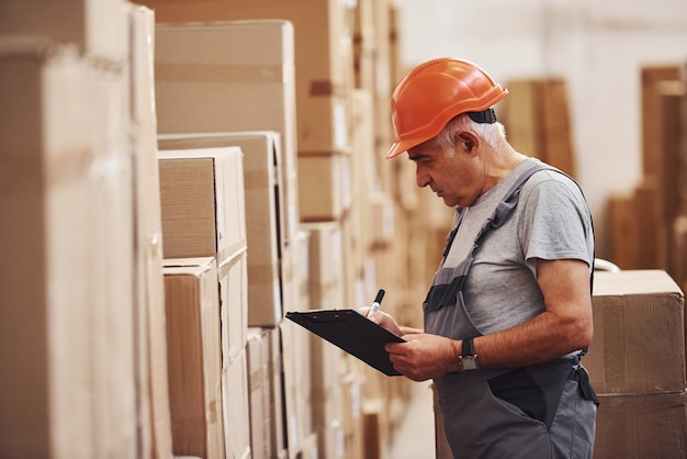 Senior storage worker in uniform and notepad in hands checks production.