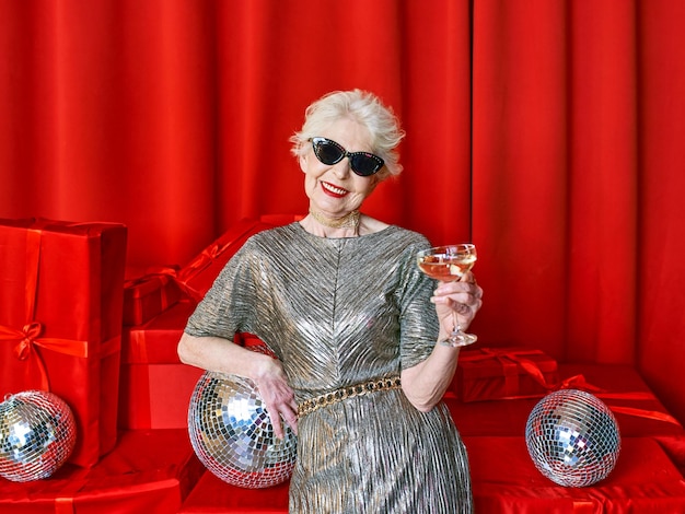 Senior stylish elegant woman with glass of sparkling wine on\
red background. party, fashion