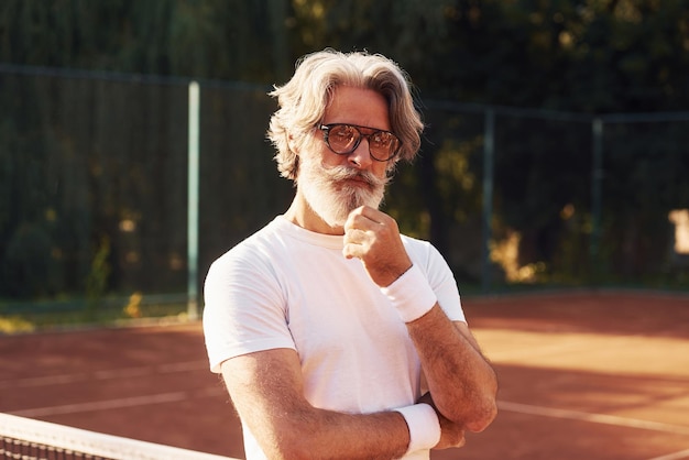 Senior stylish man in eyewear white shirt and black sportive shorts on tennis court