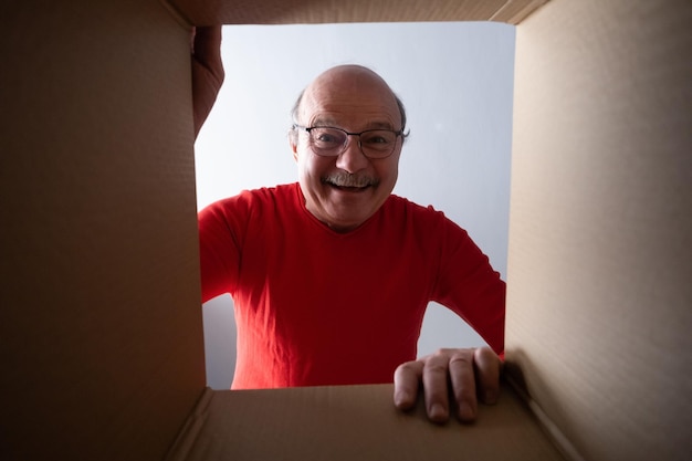Старший удивленный мужчина распаковывает, открывает картонную коробку и заглядывает внутрь.