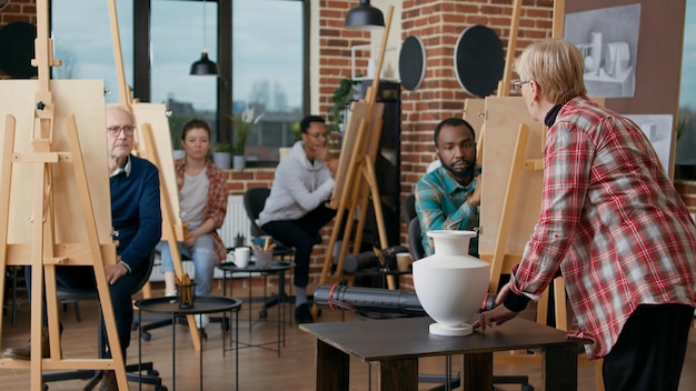 Foto insegnante senior che spiega la tecnica di disegno a un gruppo eterogeneo di studenti in classe d'arte. persone che usano matita e tela sul cavalletto per sviluppare nuove abilità artistiche e disegnare un modello di vaso sul tavolo.