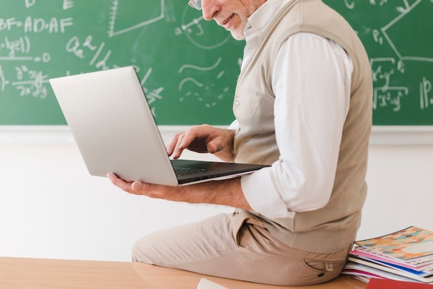 Photo senior teacher sitting on desk and surfing on laptop