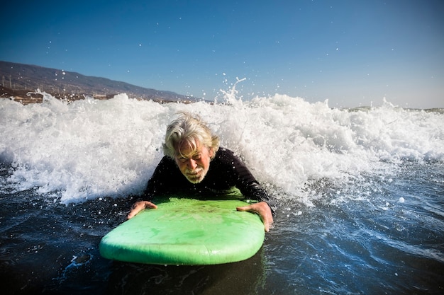 Senior trying to surf a wave on the sea at the beach alone with black wetsuit and green surftable - vacation at the sea and active retired man