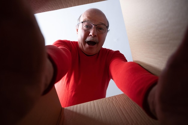 Foto senior verraste man in glazen uitpakken, kartonnen doos openen en naar binnen kijken. het pakket, levering, verrassing, cadeau lifestyle concept.