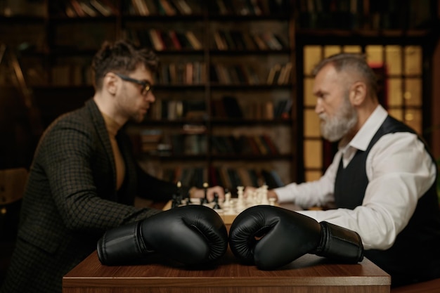 Senior versus young adult man chess battle with selective focus on boxer gloves. Business ideas, competition and strategy challenge concept