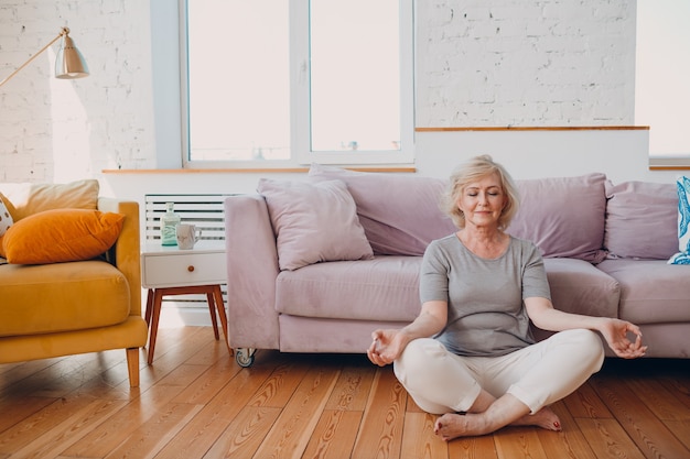 Senior volwassen lachende vrouw beoefenen van yoga thuis woonkamer. Oudere ontspannen vrouw zit in lotushouding en mediteert zen zoals in de buurt van de bank