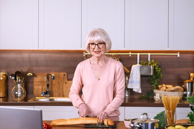 senior vrolijke vrouw kookt in de moderne keuken met een laptop