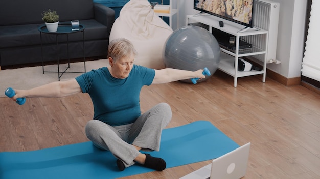 Senior vrouw die dumbbells gebruikt om gewichten op te heffen voor laptop, kijkend naar online video van trainingsles. gepensioneerde die lichaamsbeweging doet terwijl hij een trainingsprogramma volgt met een coach.