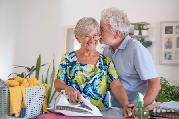 Senior vrouw die lacht terwijl ze thuis op de strijkplank strijkt terwijl haar man haar teder blanke bejaarde echtpaar met pensioen kust