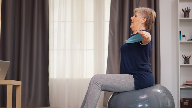 Senior vrouw die sport doet op stabiliteitsbal met weerstandsband Gepensioneerde oude vrouw die fitness uitrekt en een gezonde levensstijl leeft bij pensionering, training thuis oefening