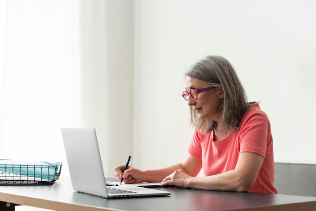 Senior vrouw die thuis studeert terwijl ze een laptop gebruikt en aantekeningen maakt