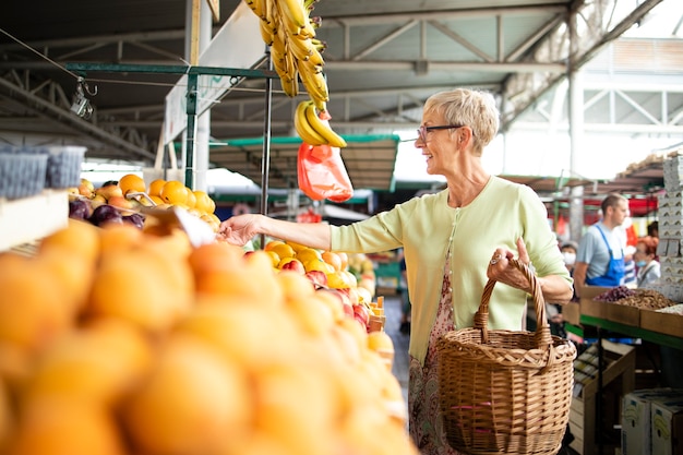 Senior vrouw die verse groenten en fruit koopt op de markt en een tas vol gezond voedsel vasthoudt