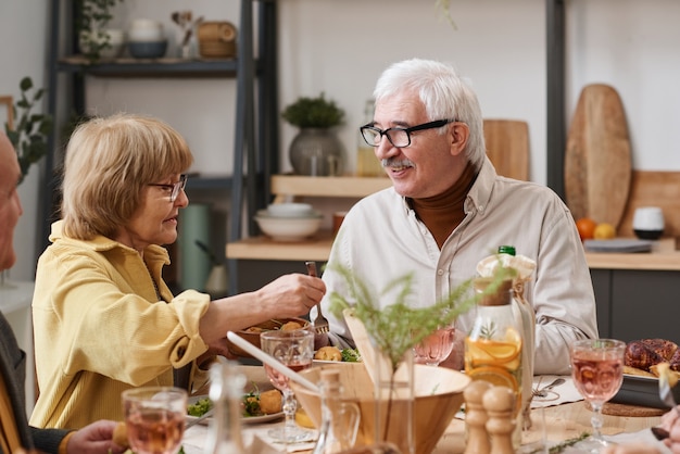 Senior vrouw die voor haar man zorgt, ze legt de maaltijd op zijn bord tijdens het familiediner