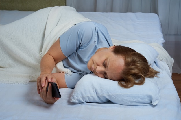 Senior vrouw in blauwe pyjama die mobiele telefoon gebruikt terwijl ze in bed ligt en lijdt aan slapeloosheid.