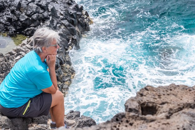 Senior vrouw in buitenexcursie op zee zittend op de klif kijkend naar de horizon over water. Pensioen levensstijl