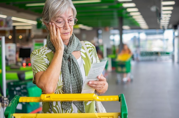 Senior vrouw in de supermarkt controleert haar boodschappenbon en kijkt bezorgd over stijgende kosten oudere dame duwt winkelwagentje consumentisme concept stijgende prijzen inflatie