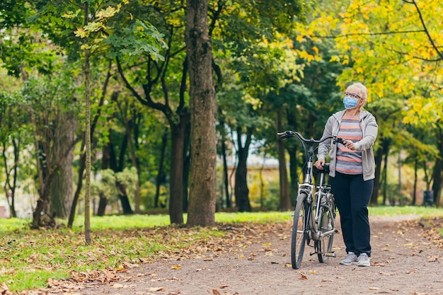 senior vrouw in een beschermend masker op haar gezicht wandelen in het park met een fiets